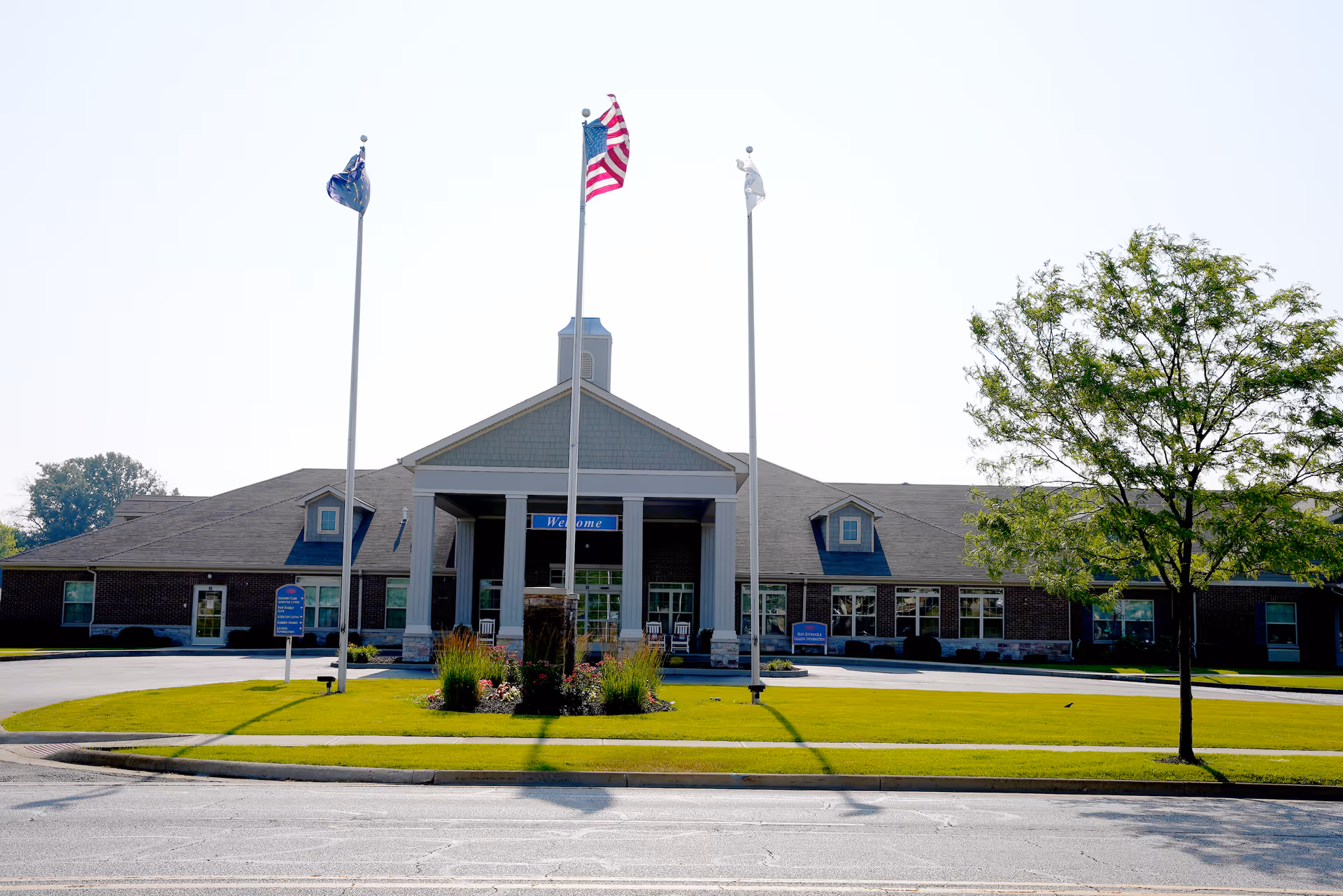 Front exterior of Aster Place senior living facility with a columned entrance, three flagpoles, manicured lawn, and circular driveway.