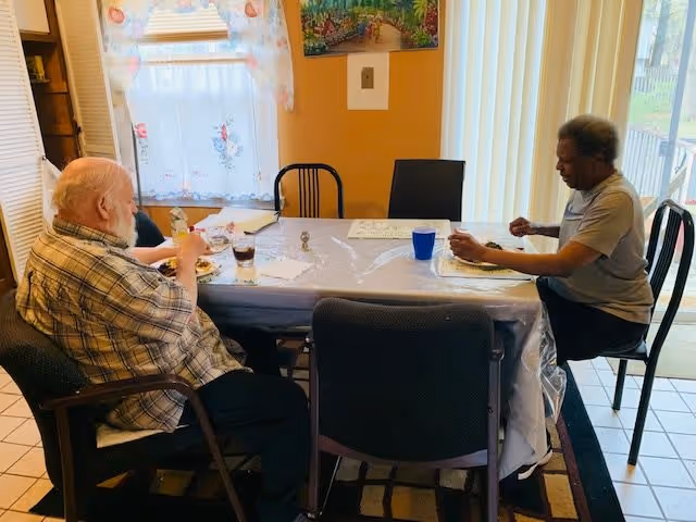Two elderly individuals sitting at a dining table covered with a plastic tablecloth, eating a meal in a room with a window covered by a floral curtain and vertical blinds. There is a painting on the wall and a glass door leading outside.