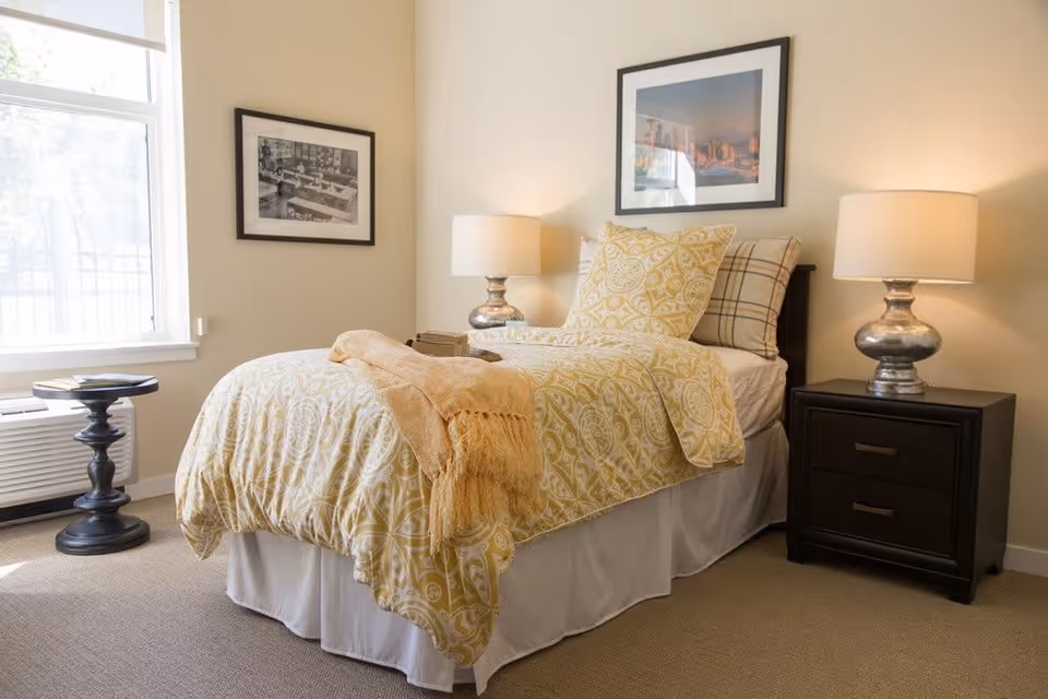 A neatly made single bed with yellow patterned bedding and pillows in a well-lit bedroom. There are two bedside tables, each with a lamp, and framed pictures hanging on the walls. A window with natural light is visible on the left side, along with a small round table near the window.