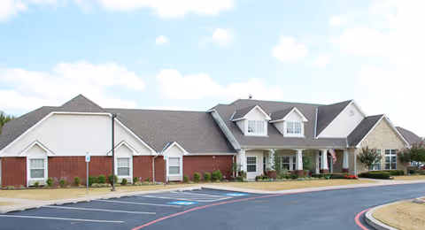 Exterior view of a single-story senior living facility building with a combination of brick and light-colored siding, multiple gabled roofs, a covered entrance with white columns, and a parking lot in front.