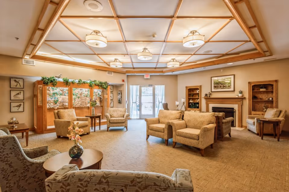 Spacious senior living lounge with armchairs, coffee tables, a fireplace, and decorative ceiling lighting.