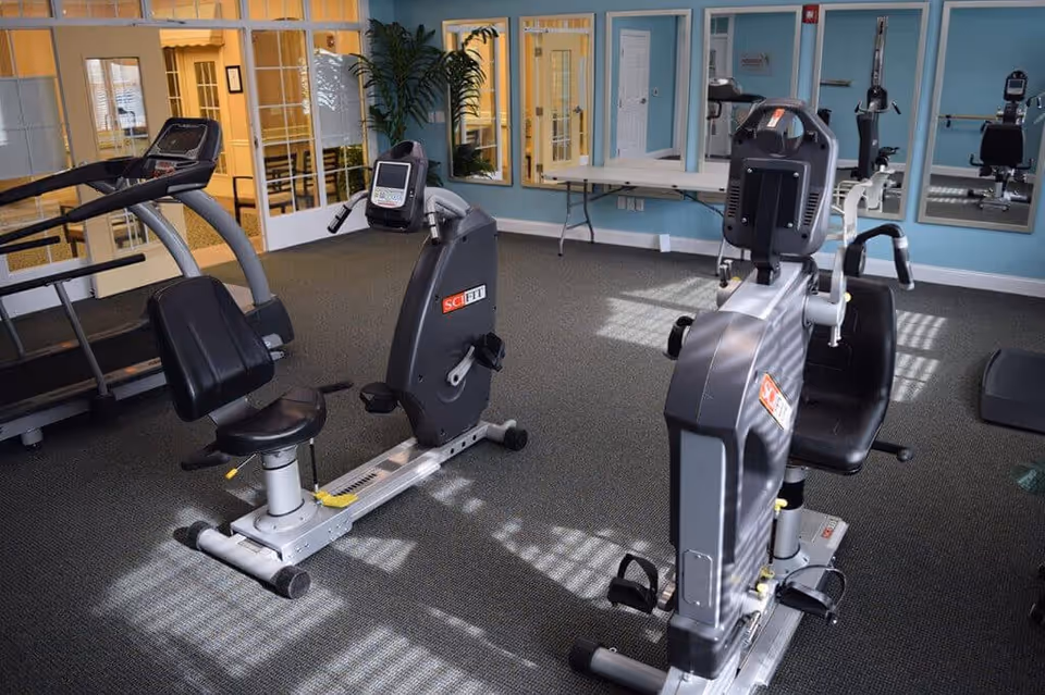 Interior view of a fitness room with exercise equipment including two recumbent stationary bikes and a treadmill. The room has blue walls, large mirrors, a potted plant, and a folding table against the wall. Sunlight streams through windows, casting shadows on the carpeted floor.