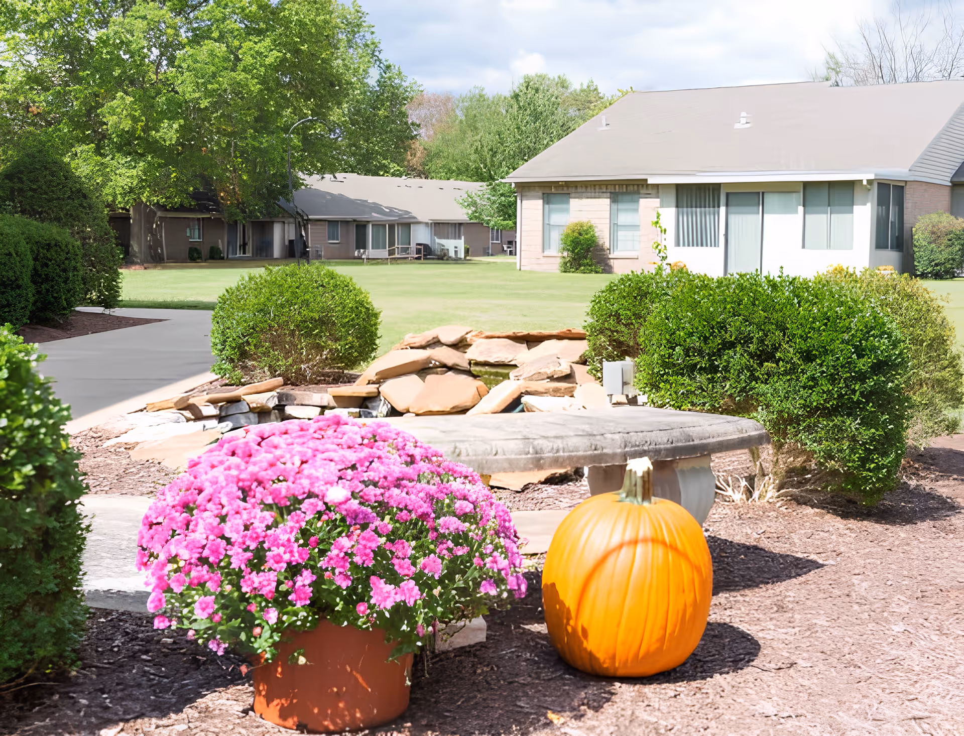 Outdoor garden area at Christian Health Center Hopkinsville featuring a large pot of pink flowers and a pumpkin in the foreground, with trimmed bushes, a stone water feature, and residential buildings in the background under a partly cloudy sky.