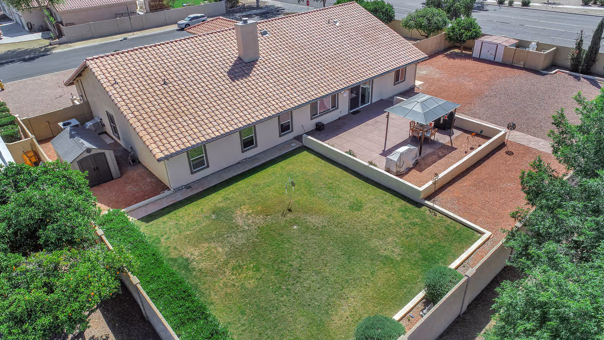 Aerial view of a single-story building with a tiled roof, surrounded by a fenced yard. The yard includes a green grassy area and a separate patio space with outdoor furniture and a canopy. There are trees and shrubs along the fence, and a small shed is visible near the building. The property is adjacent to a street with parked cars.