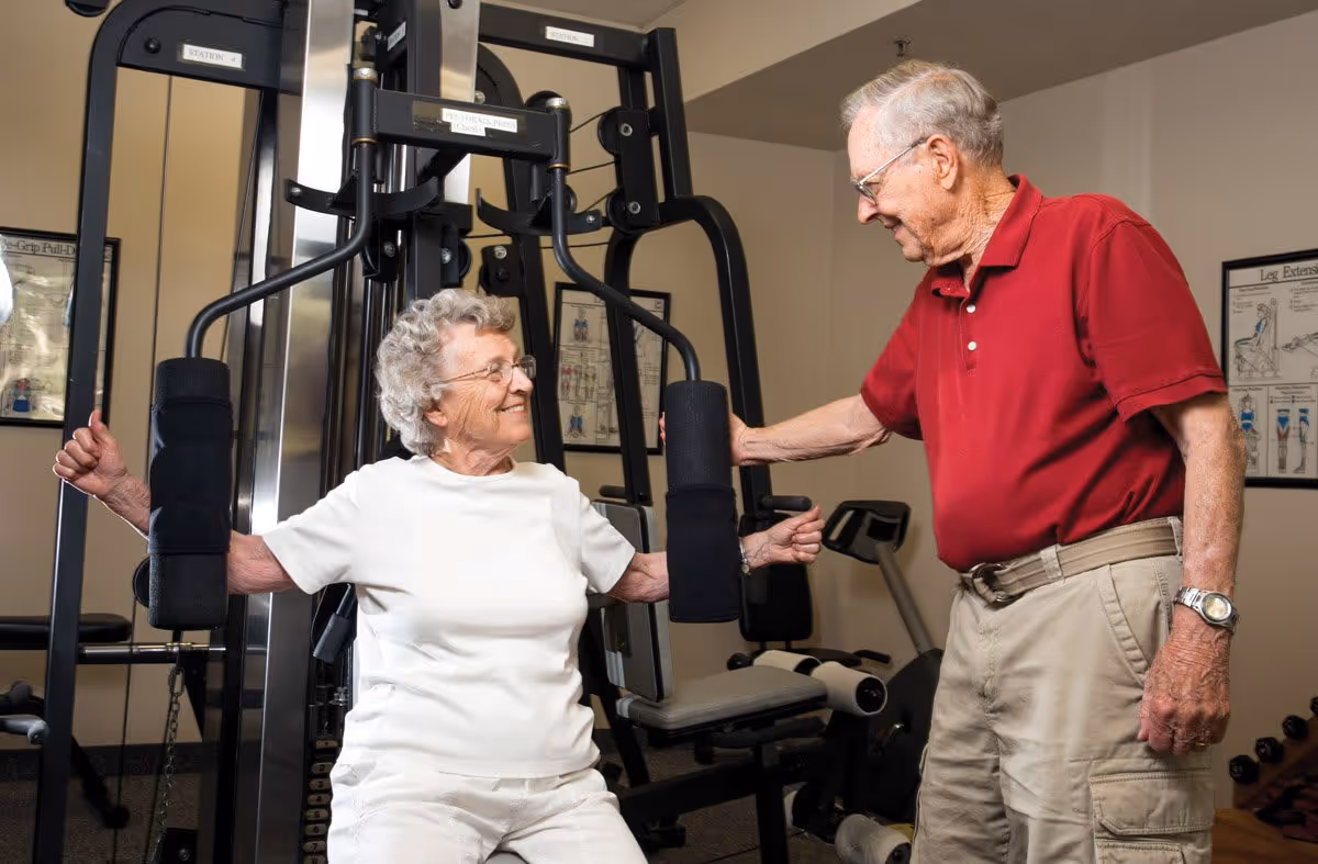 An elderly woman in a white shirt is using a chest press exercise machine in a gym, while an elderly man in a red polo shirt and beige pants stands beside her, smiling and offering support. The room contains other exercise equipment and instructional posters on the walls.