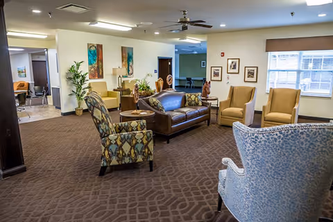 Spacious communal living room with sofas and upholstered armchairs arranged around coffee tables, decorative artwork, plants and a large window.