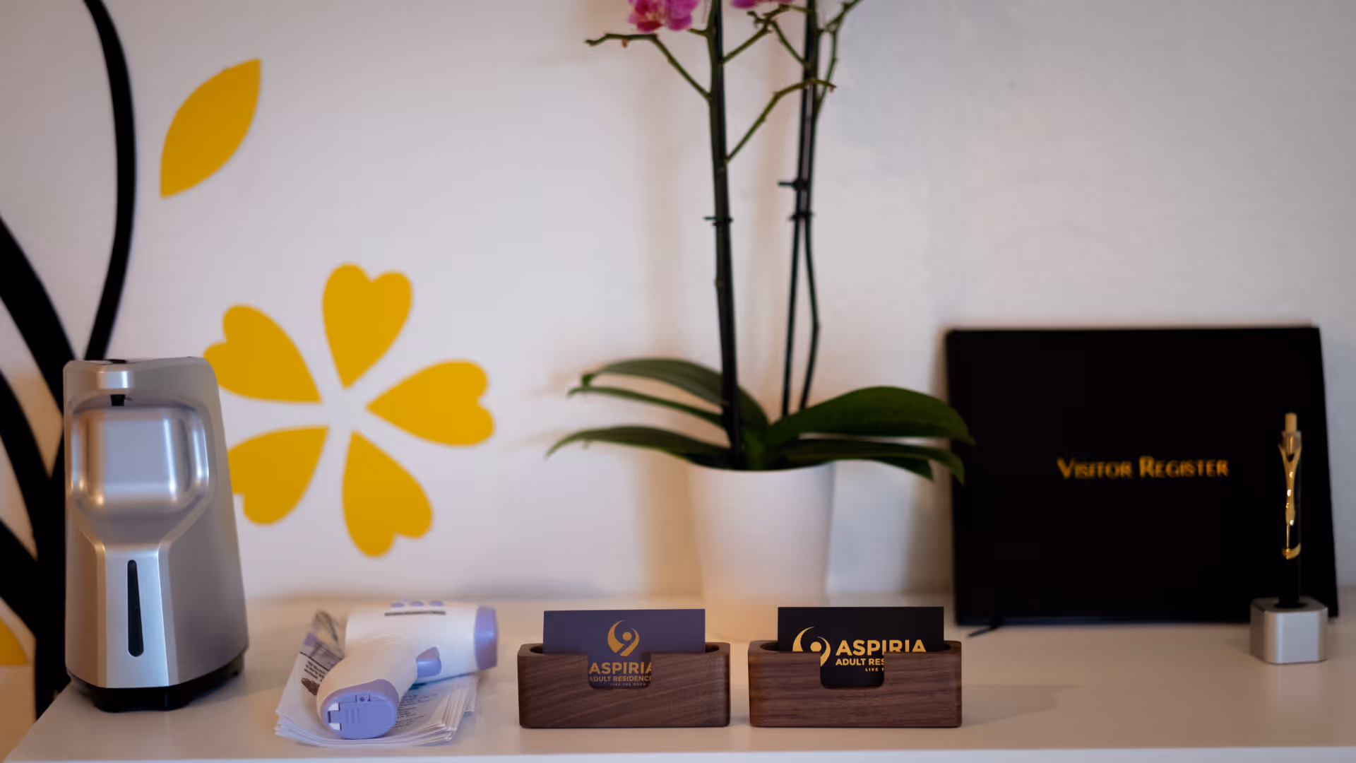 A white countertop with a silver automatic hand sanitizer dispenser, a white and purple infrared thermometer resting on papers, two wooden business card holders with Aspira Adult Residences cards, a white potted orchid plant with pink flowers, a black visitor register book, and a small pen holder with a gold pen. The background wall has a yellow floral design.