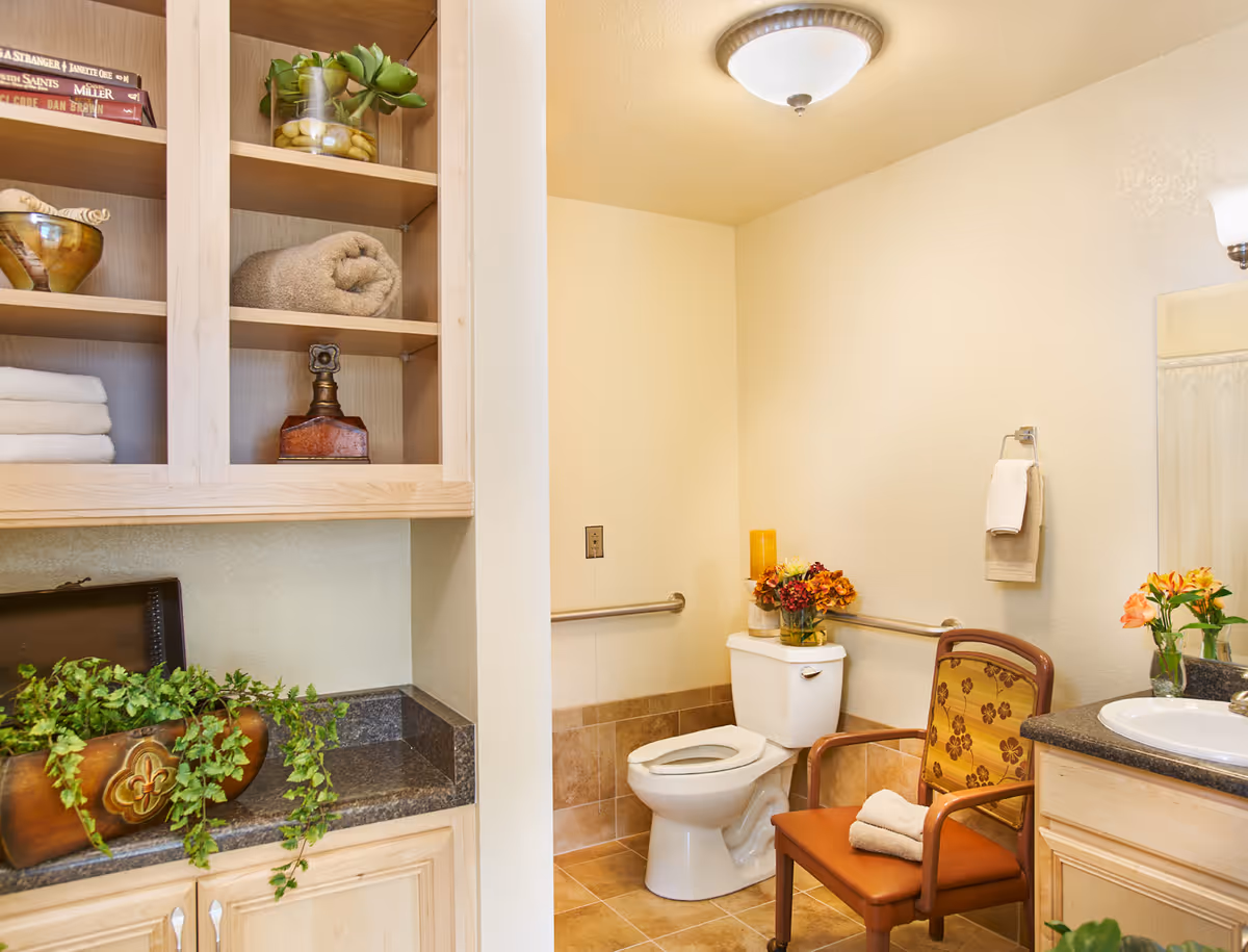 A well-lit bathroom with a white toilet, a wooden chair with a floral cushion and a folded towel on it, a countertop with a sink, and a cabinet with shelves holding towels, books, and decorative items. There are flowers placed on the toilet tank and countertop, and a towel hanging on the wall.
