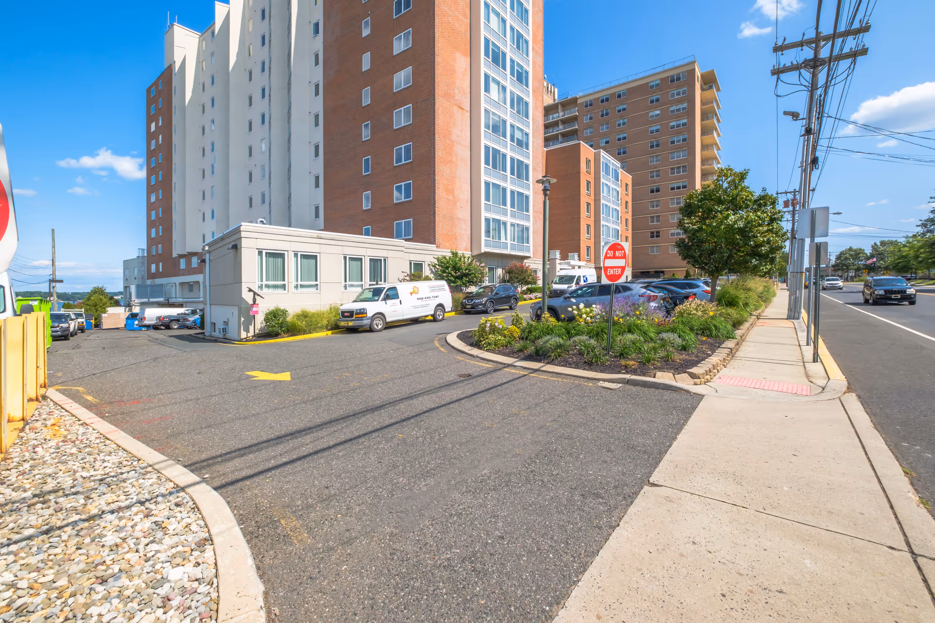 Multi-story brick-and-glass senior living building with a curved driveway, parked cars, landscaping and a sidewalk along a street.