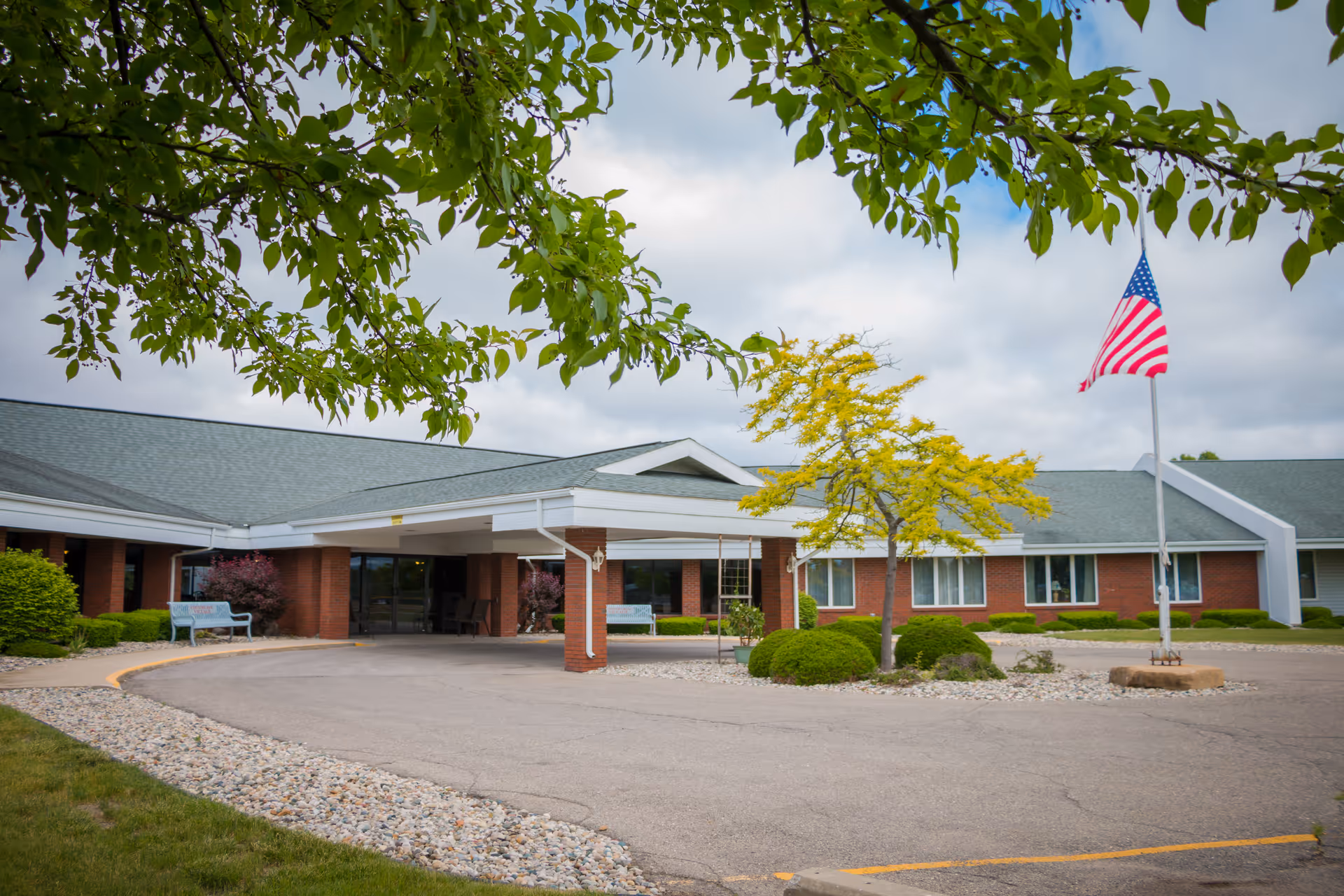 Exterior view of a single-story senior living facility building with a covered entrance, an American flag on a flagpole, trimmed bushes, a small tree with yellow leaves, and a cloudy sky. The building has a brick facade and a green roof.