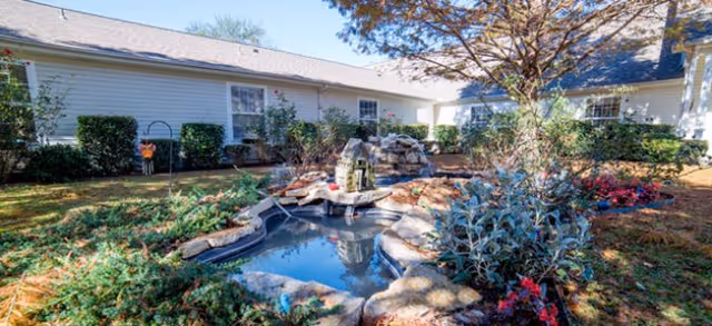 Outdoor garden area at Brookdale Tyler East featuring a small pond with a rock waterfall, surrounded by various plants, shrubs, and flowers. The garden is enclosed by a single-story building with white siding and multiple windows. A tree with bare branches is visible near the pond.