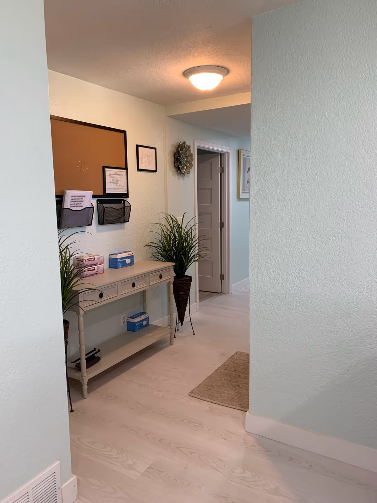 A hallway in a senior living facility with light-colored wooden flooring and pale green walls. There is a small beige console table with drawers against the left wall, holding boxes of masks and gloves. Two potted plants are placed on either side of the table. Above the table, there is a bulletin board with some papers and certificates pinned to it. A ceiling light fixture illuminates the area, and an open door leads to another room in the background.