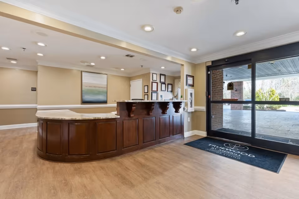 Reception area with a curved wooden front desk and marble countertop, beige walls with a framed painting, recessed ceiling lights, and a glass door entrance with a black floor mat that reads 'Truewood by Merrill Senior Living'.