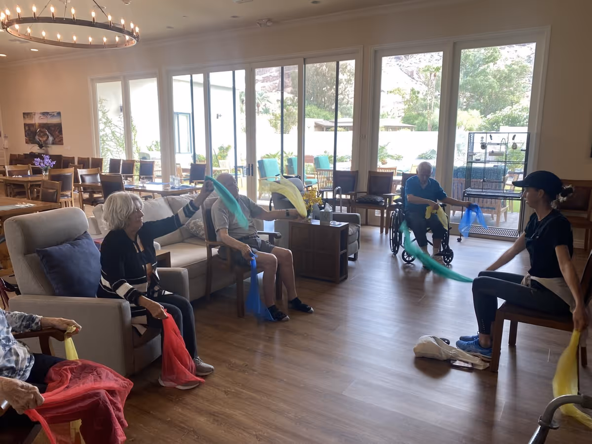 Seniors and a staff member seated in a bright communal lounge waving colorful scarves during a group activity.