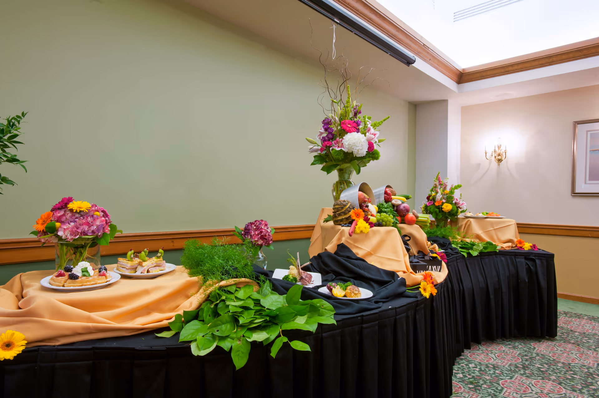 A buffet table set up with plates of food, including sandwiches and desserts, decorated with colorful flower arrangements and greenery in a room with green walls and patterned carpet.