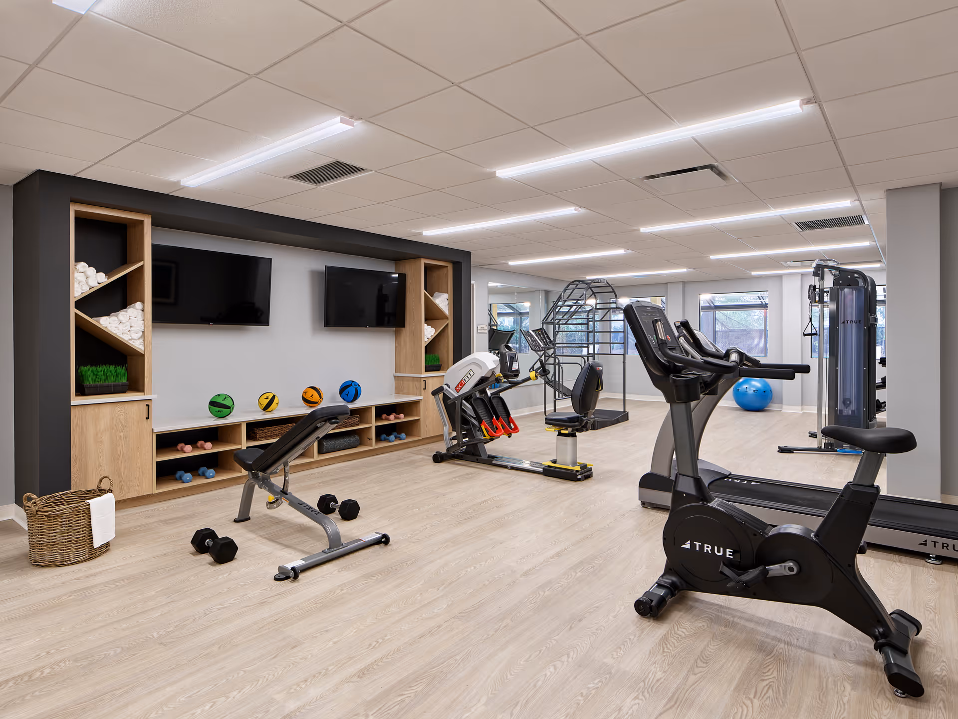 A modern fitness room with exercise equipment including a stationary bike, treadmill, rowing machine, and weight bench. The room has light wood flooring, two wall-mounted TVs, shelves with rolled towels, medicine balls, and dumbbells. Large windows and mirrors brighten the space.