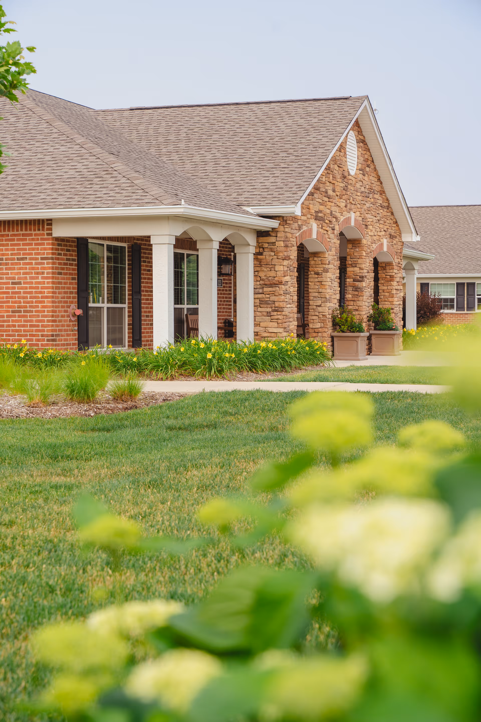 Exterior view of a senior living facility building with a combination of brick and stone facade, white columns supporting a covered porch, large windows, and well-maintained green lawn and landscaping with yellow flowers in the foreground.