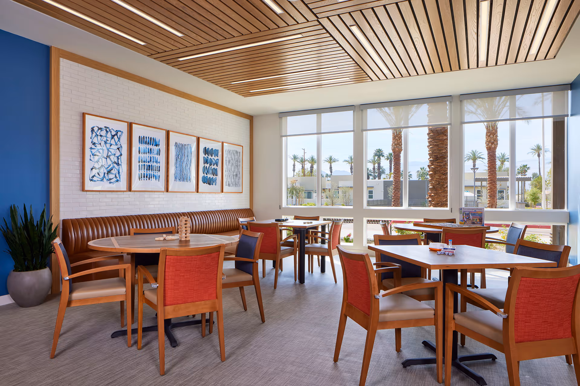 A bright and modern dining area with several wooden tables and chairs, featuring red and beige upholstery. There is a long brown cushioned bench along a white brick wall adorned with five framed abstract blue artworks. Large windows allow natural light to fill the room and offer a view of palm trees and buildings outside. The ceiling has a wooden slat design, and a potted plant is placed in the corner.