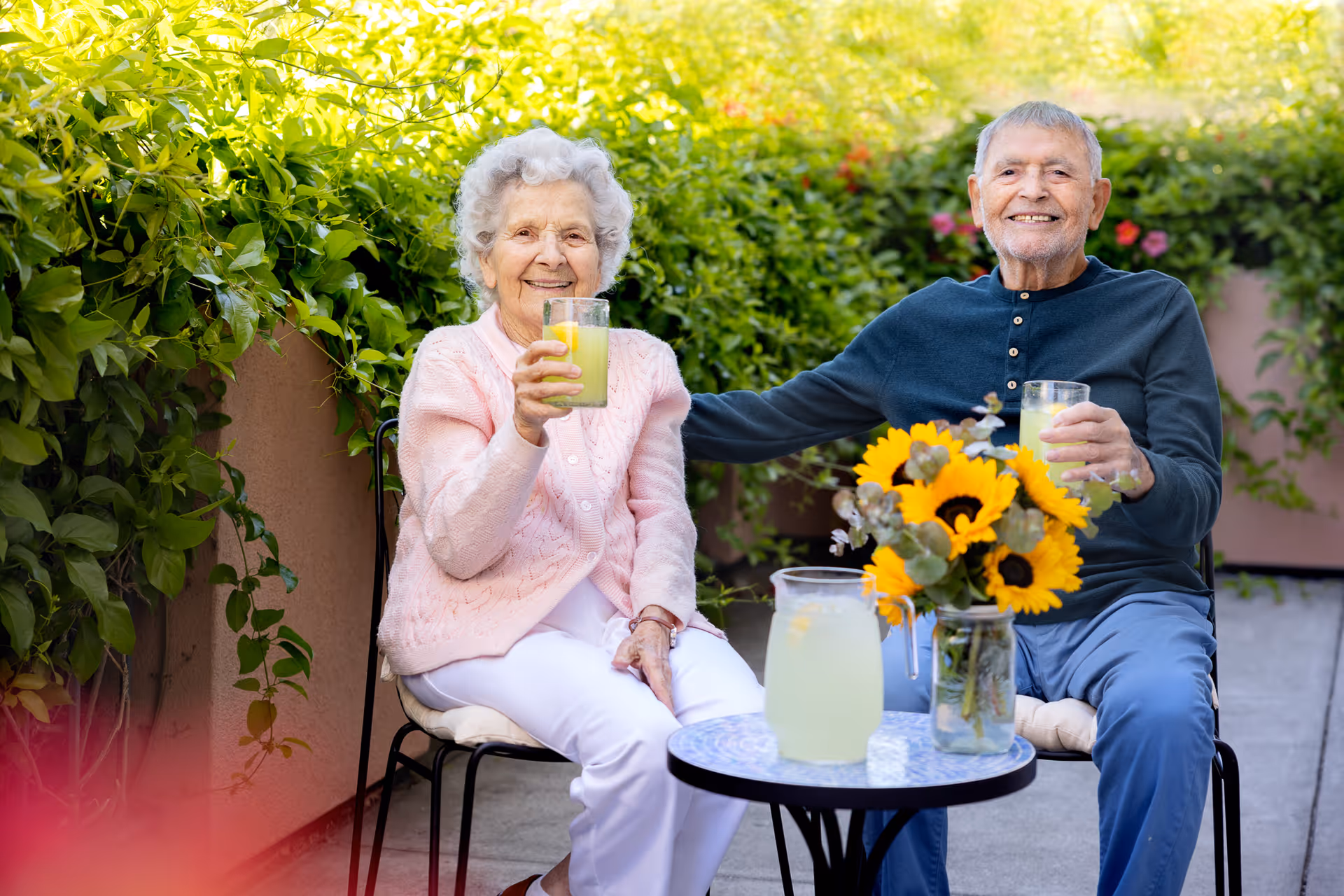 An elderly woman and man sitting outdoors at a small round table with a pitcher of lemonade and a vase of sunflowers. Both are smiling and holding glasses of lemonade, surrounded by green foliage.