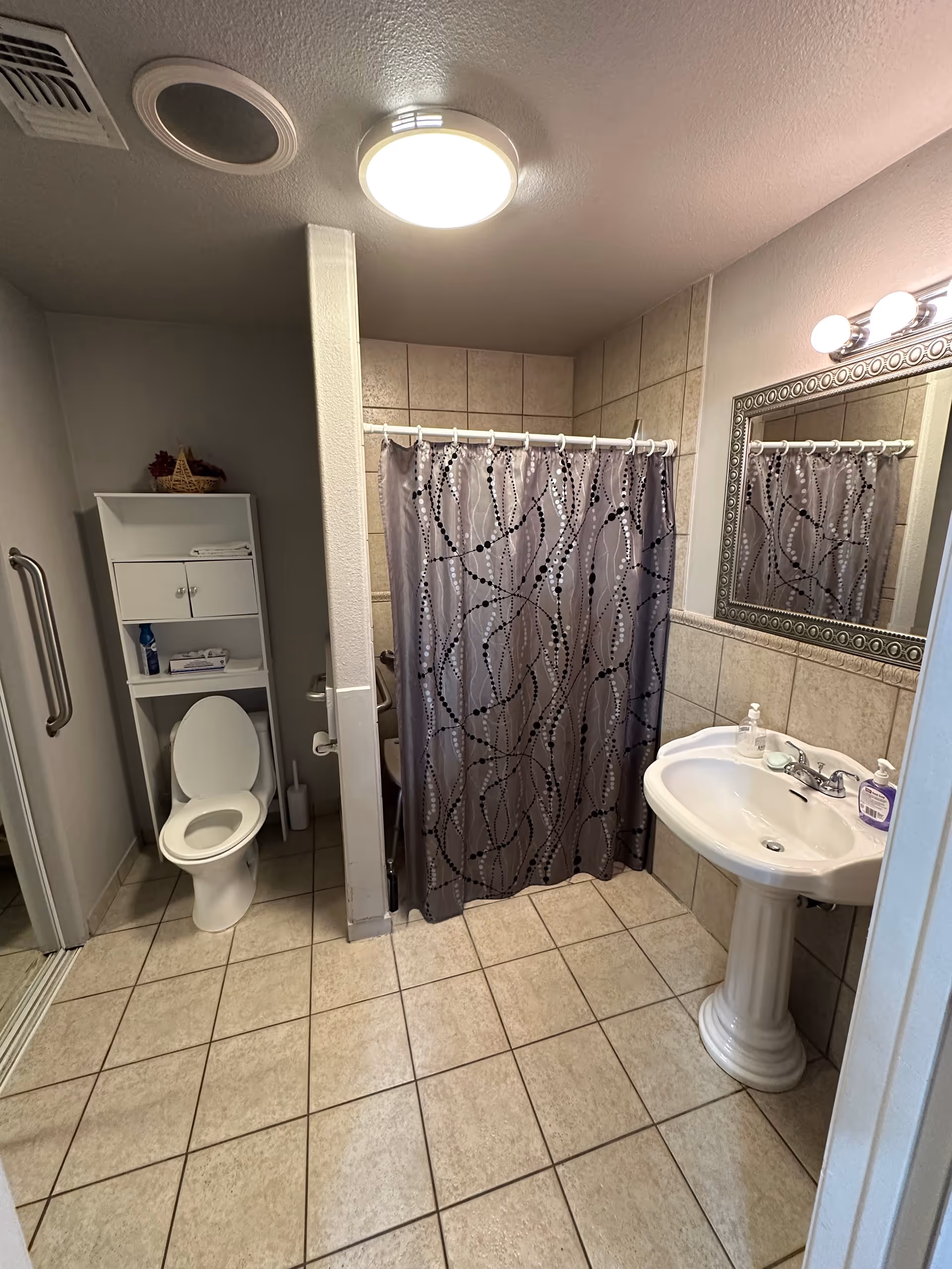 Tiled bathroom with a toilet and shelving unit, pedestal sink and mirror, and a shower with a patterned curtain.
