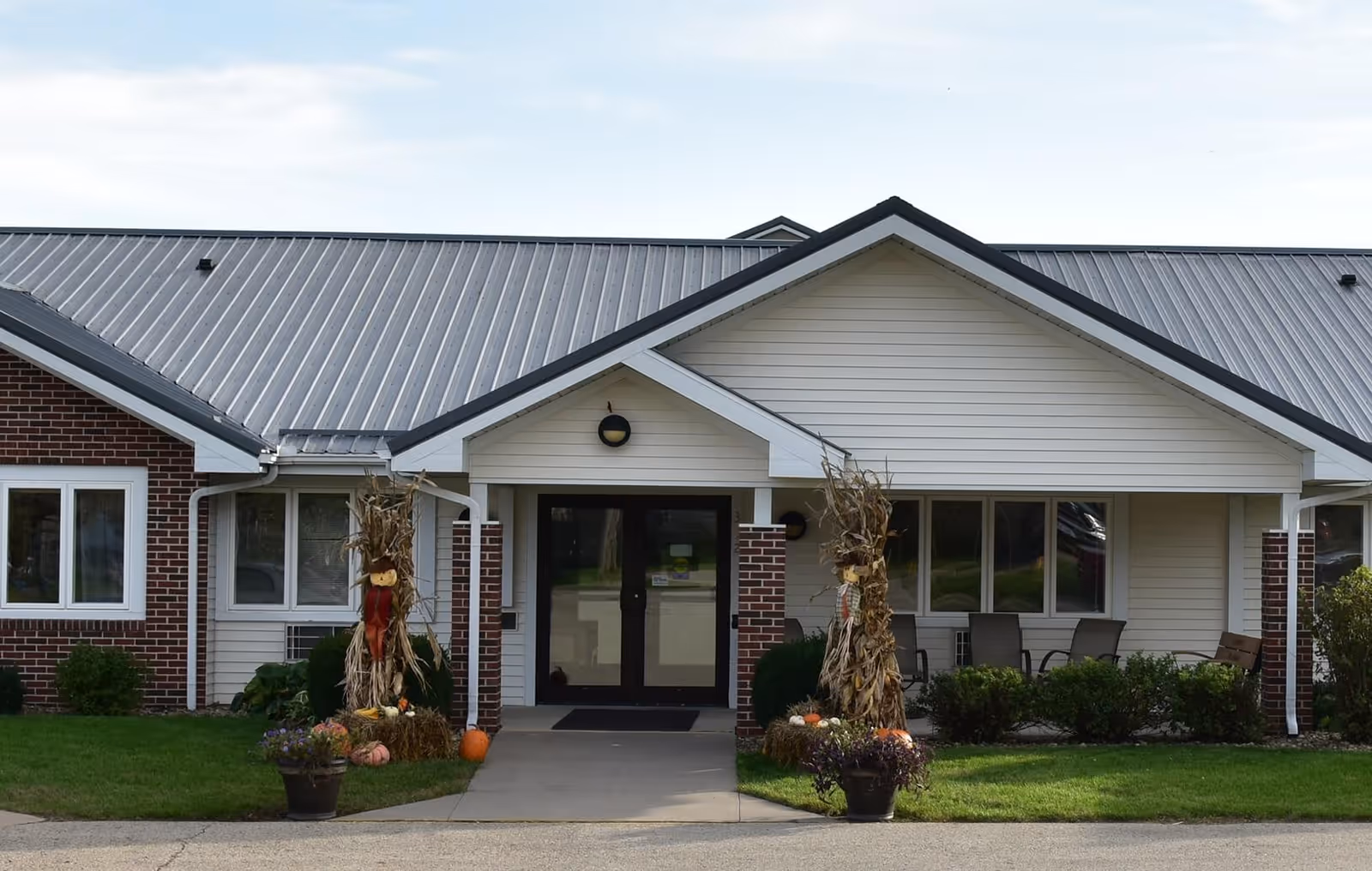 Front entrance of a single-story brick-and-siding senior care building with double glass doors, porch seating, and autumn decorations.