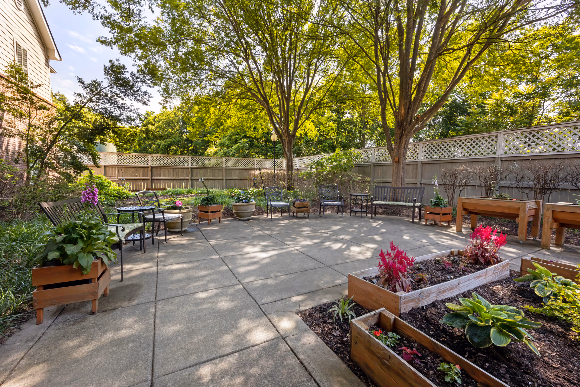 A peaceful outdoor garden area with raised wooden flower beds containing various plants and flowers. There are several metal chairs and benches arranged in a semi-circle on a concrete patio, surrounded by trees and a wooden fence with lattice top. The scene is sunlit with shadows of tree branches on the ground.