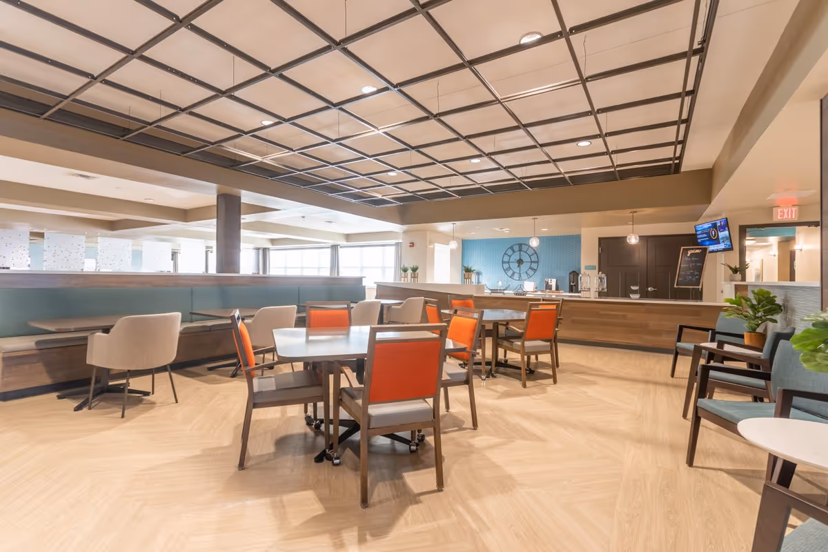 A spacious dining area with multiple tables and chairs, featuring orange and beige seating. The room has a modern ceiling design with recessed lighting, light wood flooring, and a blue accent wall with a large clock. There is a counter area with water dispensers and a small chalkboard menu, along with some potted plants and a TV mounted on the wall.