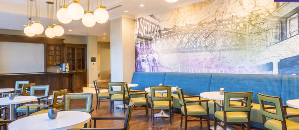 A bright dining area with round white tables and green cushioned chairs. There is a long blue cushioned bench along the wall with a large mural featuring a bridge and text. Modern globe pendant lights hang from the ceiling, and a wooden cabinet with glass doors is visible in the background.