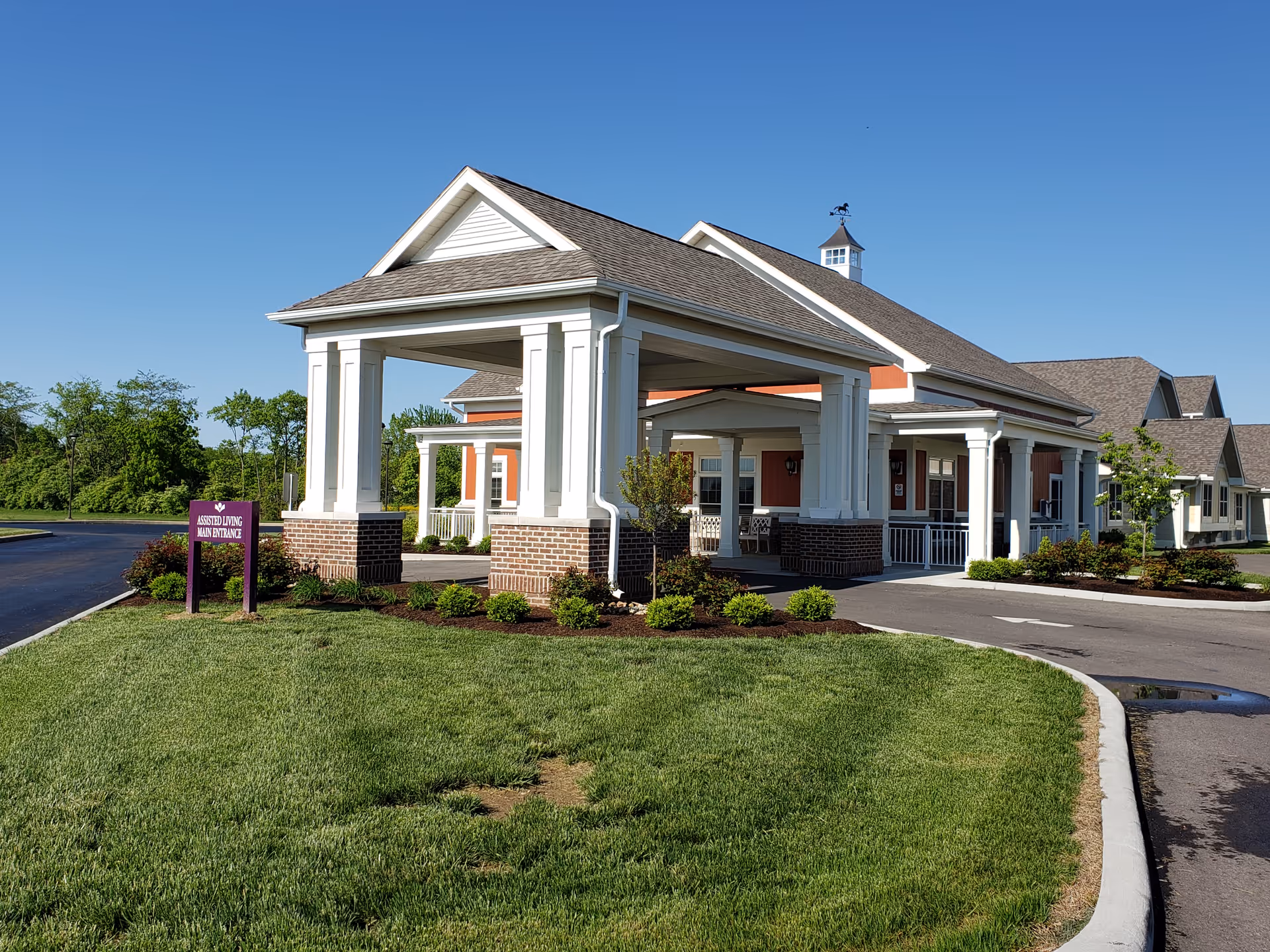 Exterior view of a senior living facility building with a covered entrance supported by white columns and brick bases. The building has a pitched roof with a small cupola on top and is surrounded by well-maintained landscaping and a paved driveway. A purple sign near the entrance reads 'Assisted Living Main Entrance'.