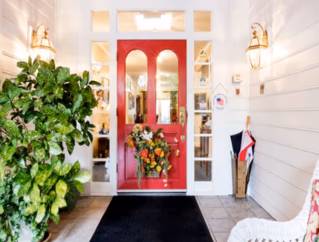 Red front door with floral arrangement and potted greenery at a white entrance.