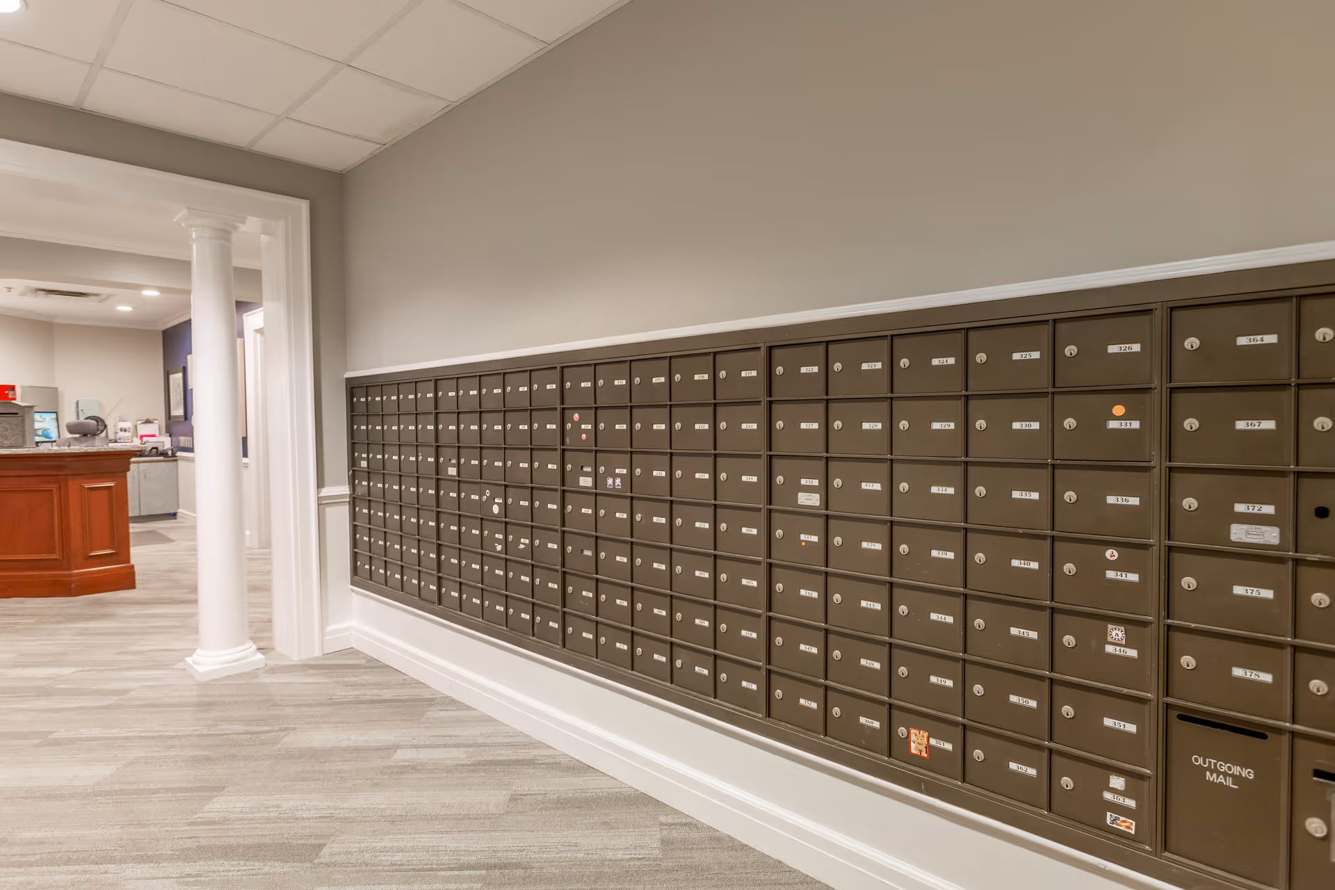 A wall of individual mailboxes in a hallway mailroom area with a reception desk visible through a doorway.