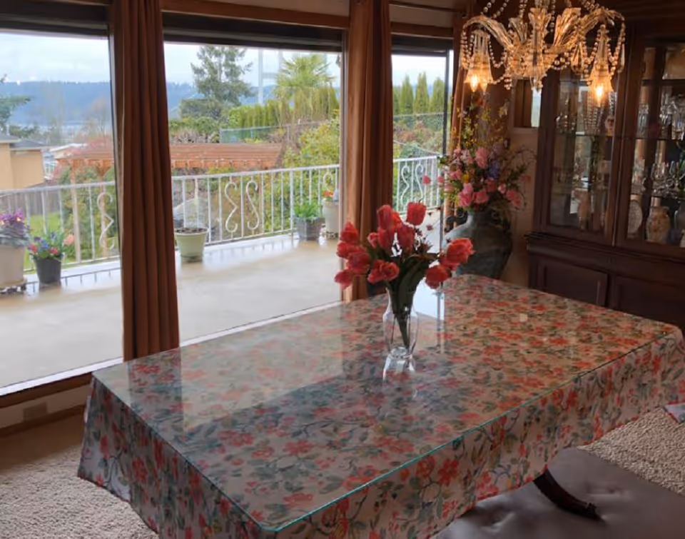 Dining room with a floral-covered table and a vase of red flowers, a chandelier and china cabinet, with large windows opening to a balcony and garden.