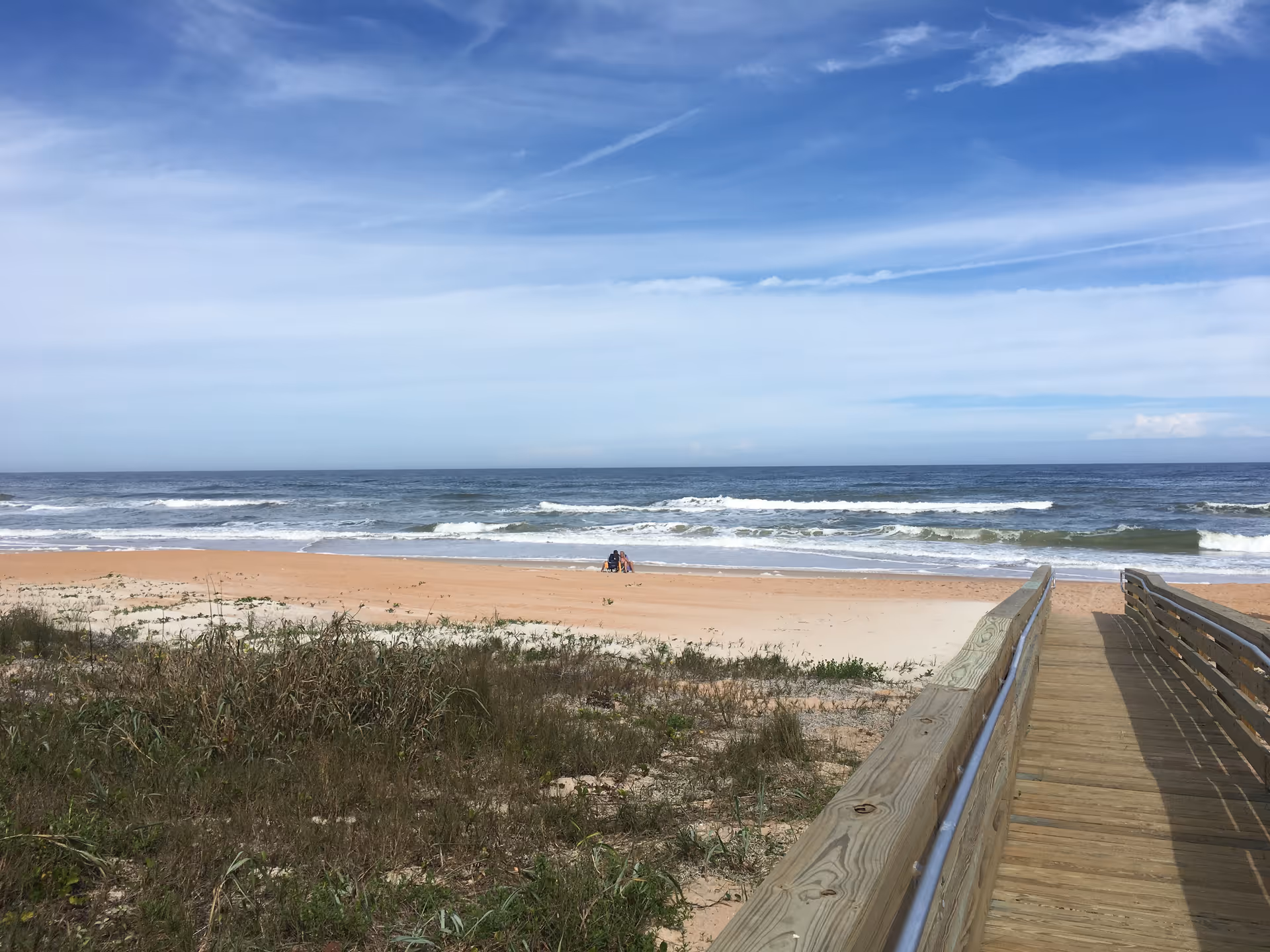 A wooden boardwalk leading to a sandy beach with ocean waves in the background under a partly cloudy blue sky. Some grass and vegetation are visible near the boardwalk and beach.