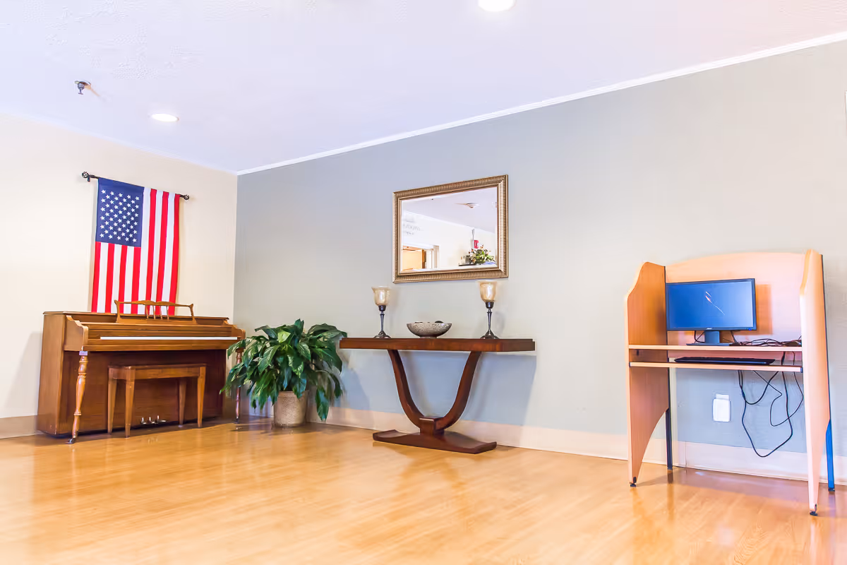 A room with a wooden piano against the left wall, an American flag hanging above it, a green potted plant next to the piano, a wooden console table with two candle holders and a decorative bowl beneath a framed mirror on the center wall, and a small wooden computer desk with a monitor and keyboard on the right side. The room has light-colored walls and a wooden floor.