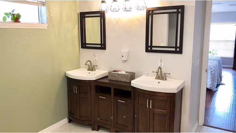 A bathroom interior featuring a double sink vanity with dark wood cabinets and two rectangular mirrors above each sink. There are three decorative light fixtures mounted on the wall above the mirrors. A small window with a potted plant is visible on the left wall, and an open doorway on the right leads to a bedroom with a bed and wooden floor.