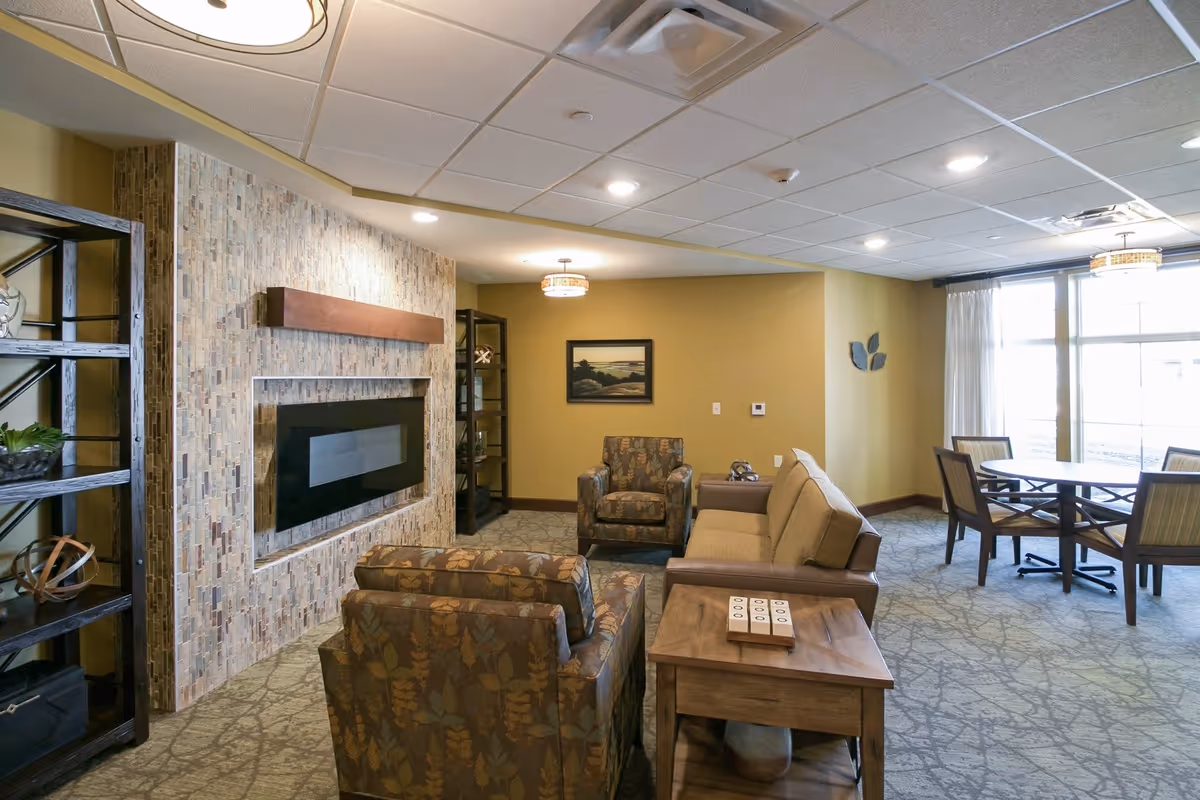 A cozy living room area in a senior living facility with patterned armchairs, a beige sofa, a wooden side table, and a modern electric fireplace set in a tiled wall. The room has carpeted floors, a large window with sheer curtains, and a round dining table with chairs near the window. Shelves with decorative items flank the fireplace, and the walls are painted a warm beige color.