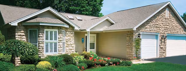 Exterior view of a single-story residential building with stone and siding facade, a three-car garage, and a well-maintained garden with shrubs and flowers in front.