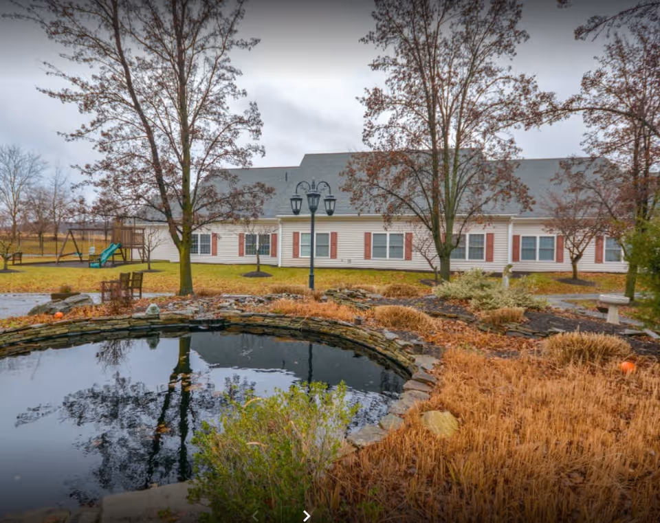 Outdoor view of St. Martha Center for Rehabilitation & Healthcare showing a small pond surrounded by stone edging and dry vegetation. There are several leafless trees, a playground with a slide, benches, and a lamppost in front of a single-story building with white siding and red shutters under a cloudy sky.
