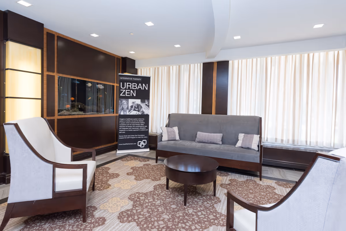 A bright and modern lounge area with a gray sofa, two white armchairs with wooden frames, a round dark wooden coffee table, and a patterned carpet. Behind the sofa are large windows with sheer curtains. On the left wall, there is a built-in fish tank and a standing banner that reads 'Integrative Therapy Urban Zen.'