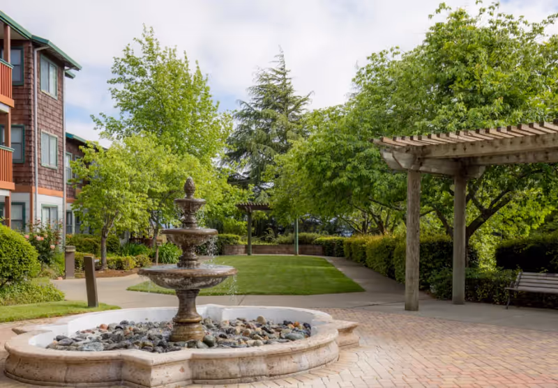 Outdoor garden area at Cogir Of Vallejo Hills featuring a three-tier stone fountain surrounded by rocks, a paved walkway, green grass, trees, bushes, a wooden pergola, and a bench. The side of a multi-story building with windows and balconies is visible on the left.