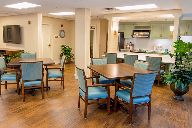 A bright and clean common area with wooden floors, several square wooden tables surrounded by blue cushioned chairs, a kitchen area with green cabinets and stainless steel appliances in the background, and potted plants adding greenery to the space.