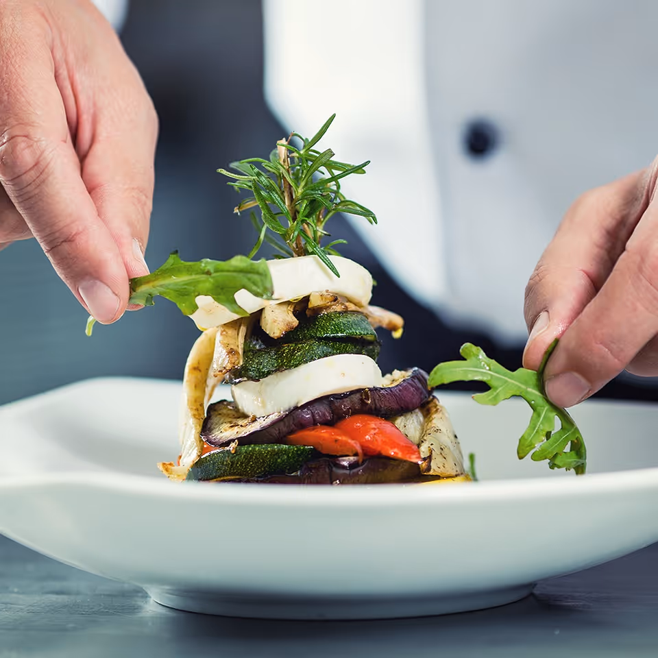 Chef's hands arranging arugula on a stacked grilled vegetable and cheese tower garnished with herbs on a white plate.