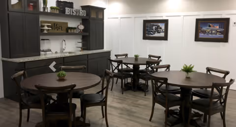 A dining area with several round wooden tables and chairs, each table decorated with a small green plant. The room features a kitchenette with dark cabinets, a granite countertop, a sink, and open shelves with decorative items. Two framed pictures hang on the wall above the tables.