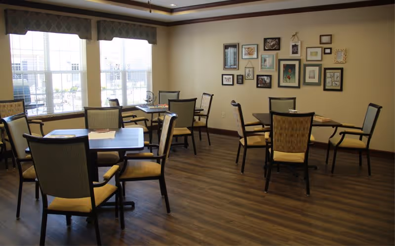 A well-lit room with several square tables and cushioned chairs arranged around them. The room has large windows with valances letting in natural light. On one wall, there is a gallery of framed pictures and artwork. The floor is wood, and the overall decor is warm and inviting.