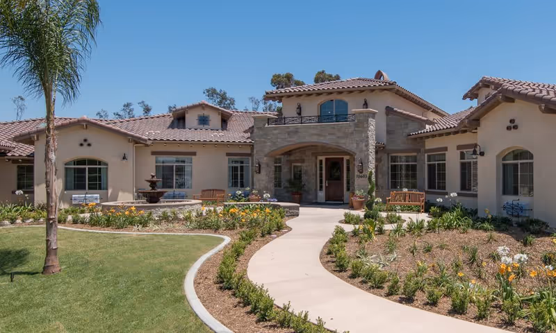 Exterior view of a single-story senior living facility building with a tiled roof, arched entrance, and landscaped garden with flowers and shrubs along a curved walkway leading to the front door under a clear blue sky.