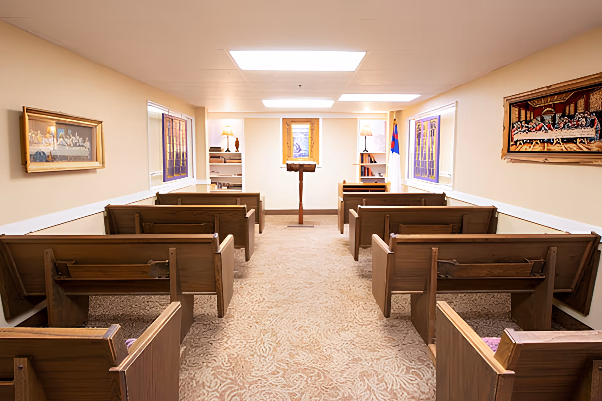Interior view of a small chapel or worship room with wooden pews arranged in two rows facing a wooden lectern. The room has beige walls, carpeted floor, and ceiling lights. There are framed religious artworks on the walls and a Christian flag near the front.