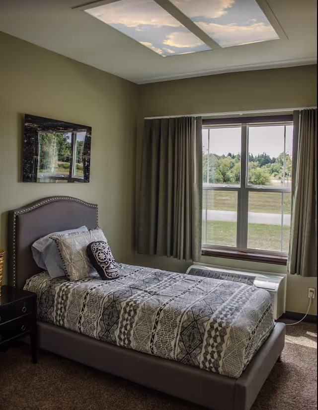 Single bedroom with a patterned twin bed, nightstand, wall art, and a window overlooking a grassy outdoor area.