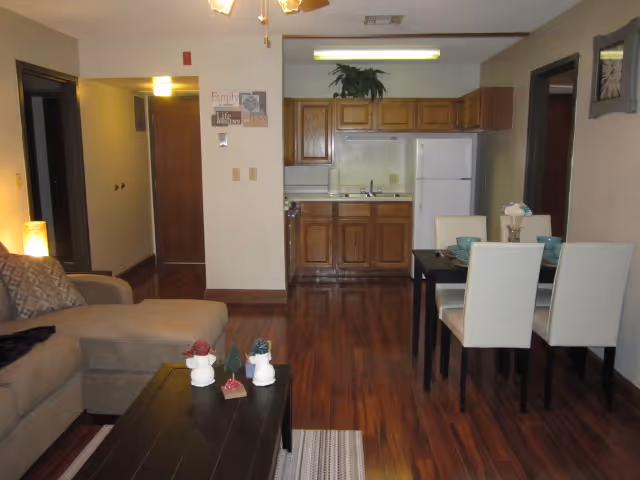 Interior view of a senior living facility apartment showing a living room with a beige sectional sofa and a dark wooden coffee table with decorative items. Adjacent to the living room is a dining area with a black dining table and four white chairs set with blue dishes. The kitchen area in the background has wooden cabinets, a white refrigerator, and a sink. The floor is hardwood, and the walls are light-colored with some framed artwork and a ceiling fan with lights.