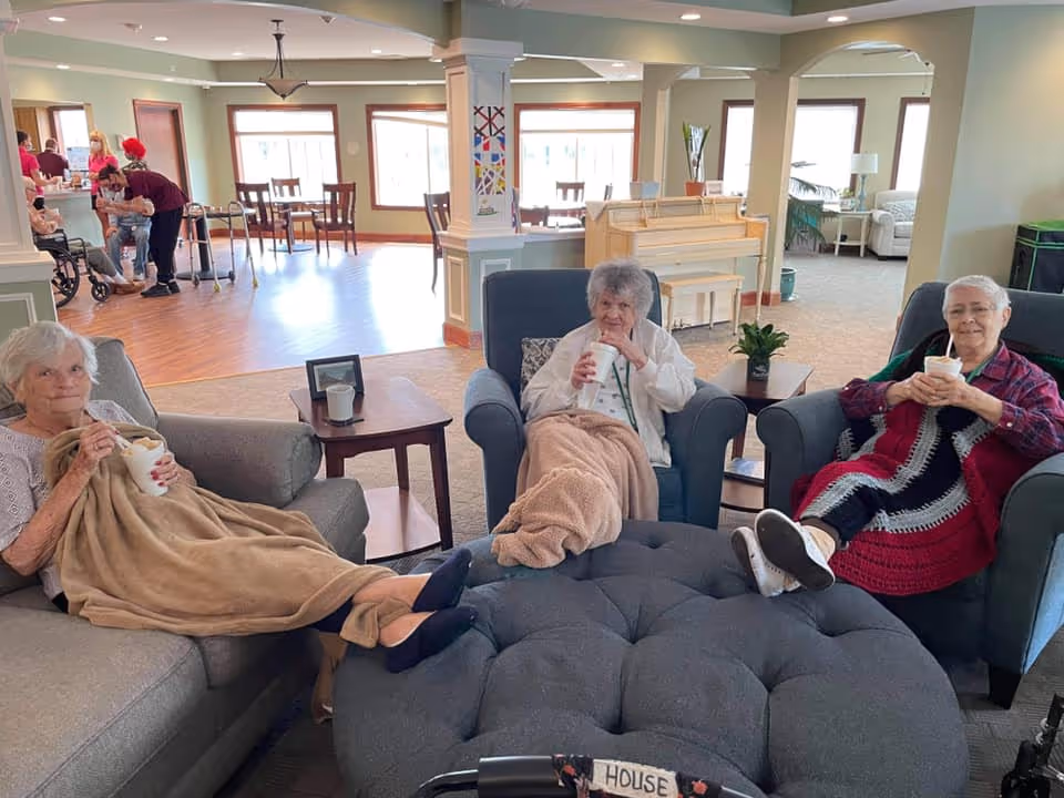 Three elderly women sitting comfortably in armchairs in a cozy living room area, each holding a drink and covered with blankets. In the background, a few staff members assist other residents near a dining area with tables and chairs, and a piano is visible against the wall.