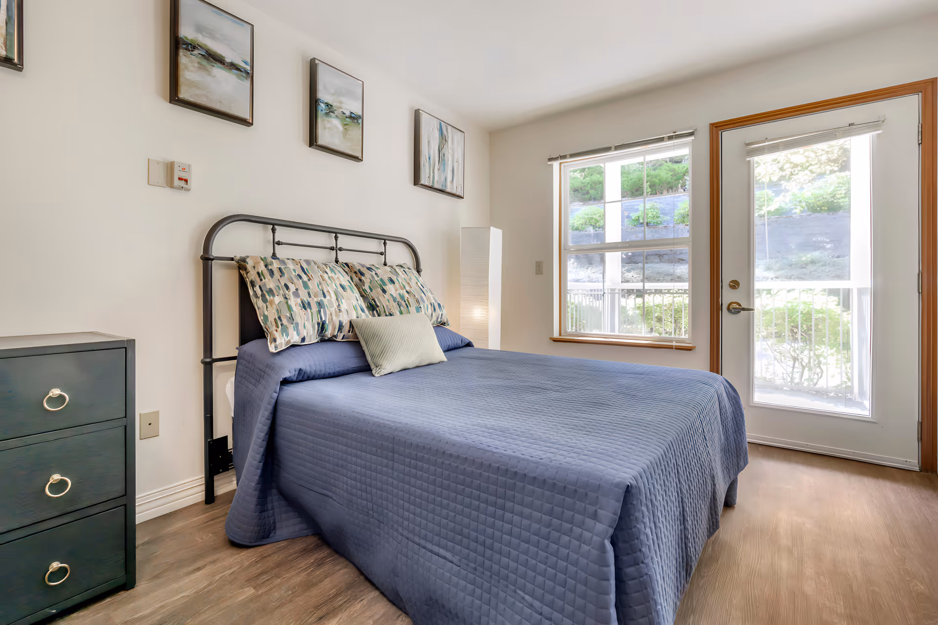 Bright bedroom with a blue-quilted bed, decorative pillows, a bedside dresser, window and glass door to the outside.